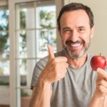 Cheerful mature man eating an apple and showing his thumb up.