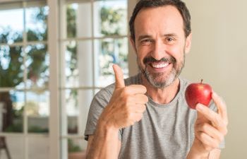 Cheerful mature man eating an apple and showing his thumb up.