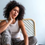 young woman with curly hair touching cheek in pain having toothache sitting on bed