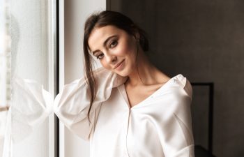 Image of gorgeous woman 30s smiling and standing near big window in living room